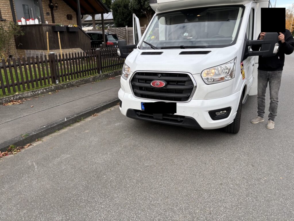 Das Bild zeigt die Vorderseite eines weißen Wohnmobils in einer Wohngegend, wobei eine Person mit verdecktem Gesicht neben dem Fahrzeug steht. Im Hintergrund sind ein Haus mit Zaun und ein weiteres Auto zu sehen.