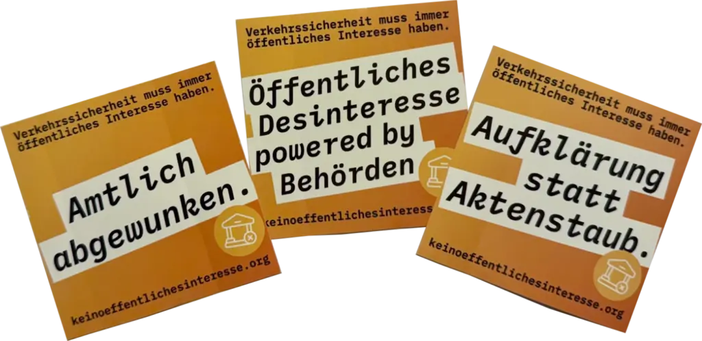 Das Bild zeigt drei orangefarbene Aufkleber auf einem weißen Blatt Papier mit der Aufschrift "Verkehrssicherheit muss immer öffentliches Interesse haben." Die Aufkleber enthalten die Slogans "Amtlich abgewunken", "Öffentliches Desinteresse powered by Behörden" und "Aufklärung statt Aktenstaub."