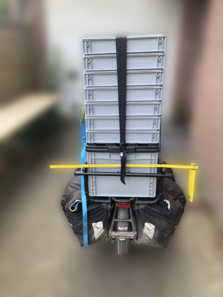 Ein Fahrrad mit mehreren grauen Kisten, die übereinander gestapelt sind, ist mit einem schwarzen Gurt gesichert. Daneben ist eine gelbe Messlatte angebracht, die als Abstandshalter dient.