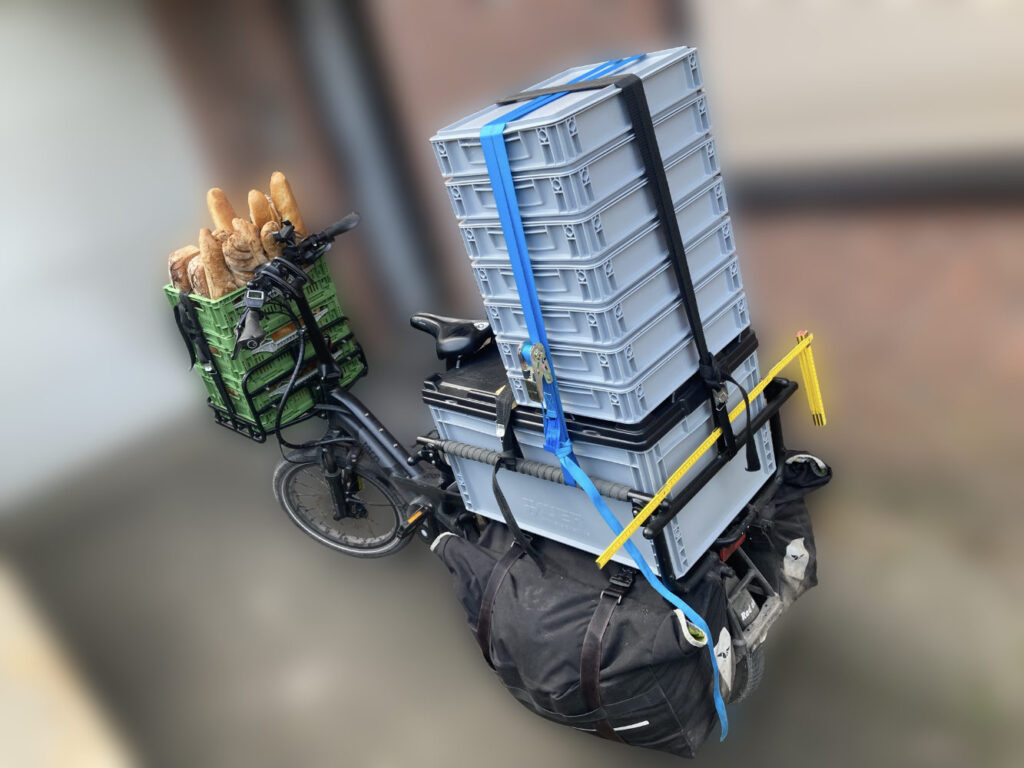 Ein Lastenrad ist vor einer roten Ziegelwand geparkt und mit mehreren Stapeln von grauen Kisten und einem grünen Korb voller Brot beladen. Die Kisten und der Korb sind mit blauen und schwarzen Gurten gesichert.